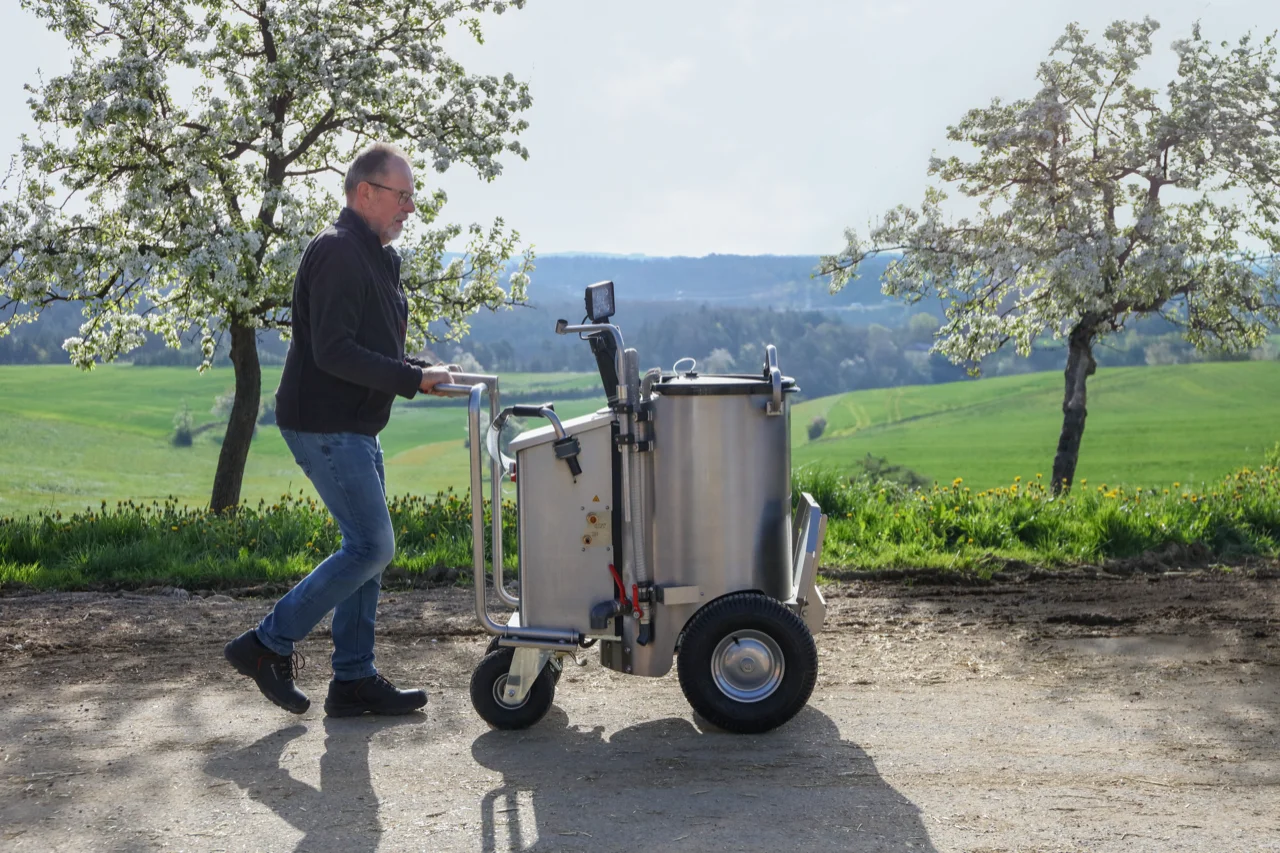 A farmer pulls the MilchMobil through an idyllic, green landscape. Surrounded by nature, he uses the device in a natural environment to feed his calves.