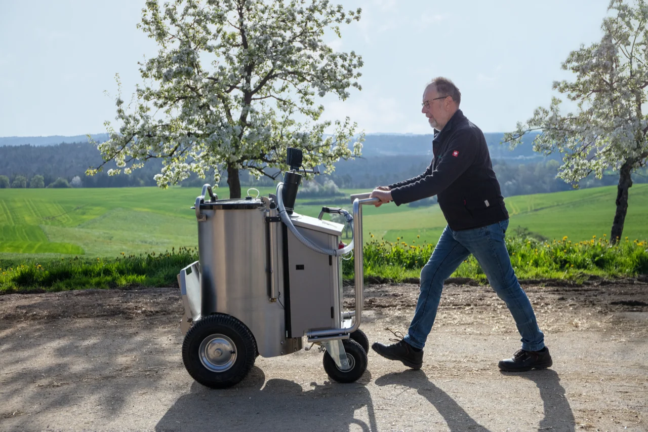 Ein Landwirt schiebt das MilchMobil durch eine natürliche Umgebung, umgeben von grüner Landschaft.